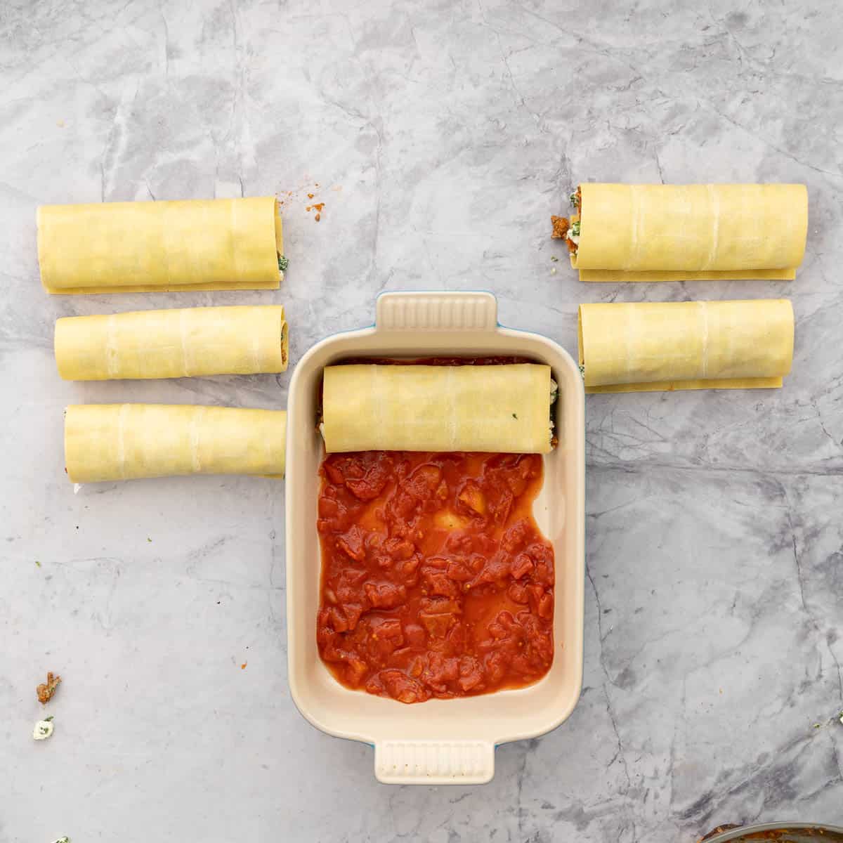 Rolled up pasta sheets next to a dish which has a layer of diced tomatoes in the bottom and one lasagna roll up placed in the on top 