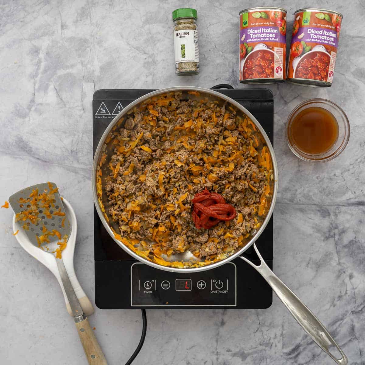 Browned mince in a pan with tomato paste added which is cooking on a bench top cooker next to two cans of diced tomatoes and a glass cup of broth and a spatula 