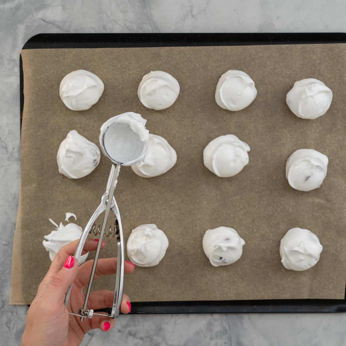 Large spoonfuls of meringue mixture on a linebacking tray with a hand above it holding a cookie scoop. 