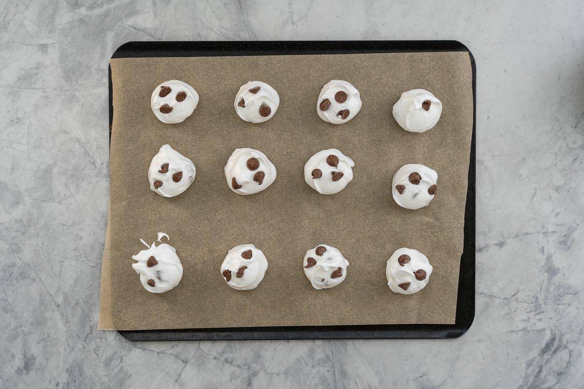 Large spoonfuls of meringue mixture on a linebacking tray with added chocolate drops added to the top each one.