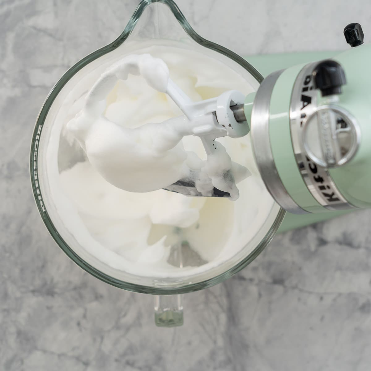 A stand up mixer sitting on the bench with whipped egg whites with the beaters lifted showing soft peaks 
