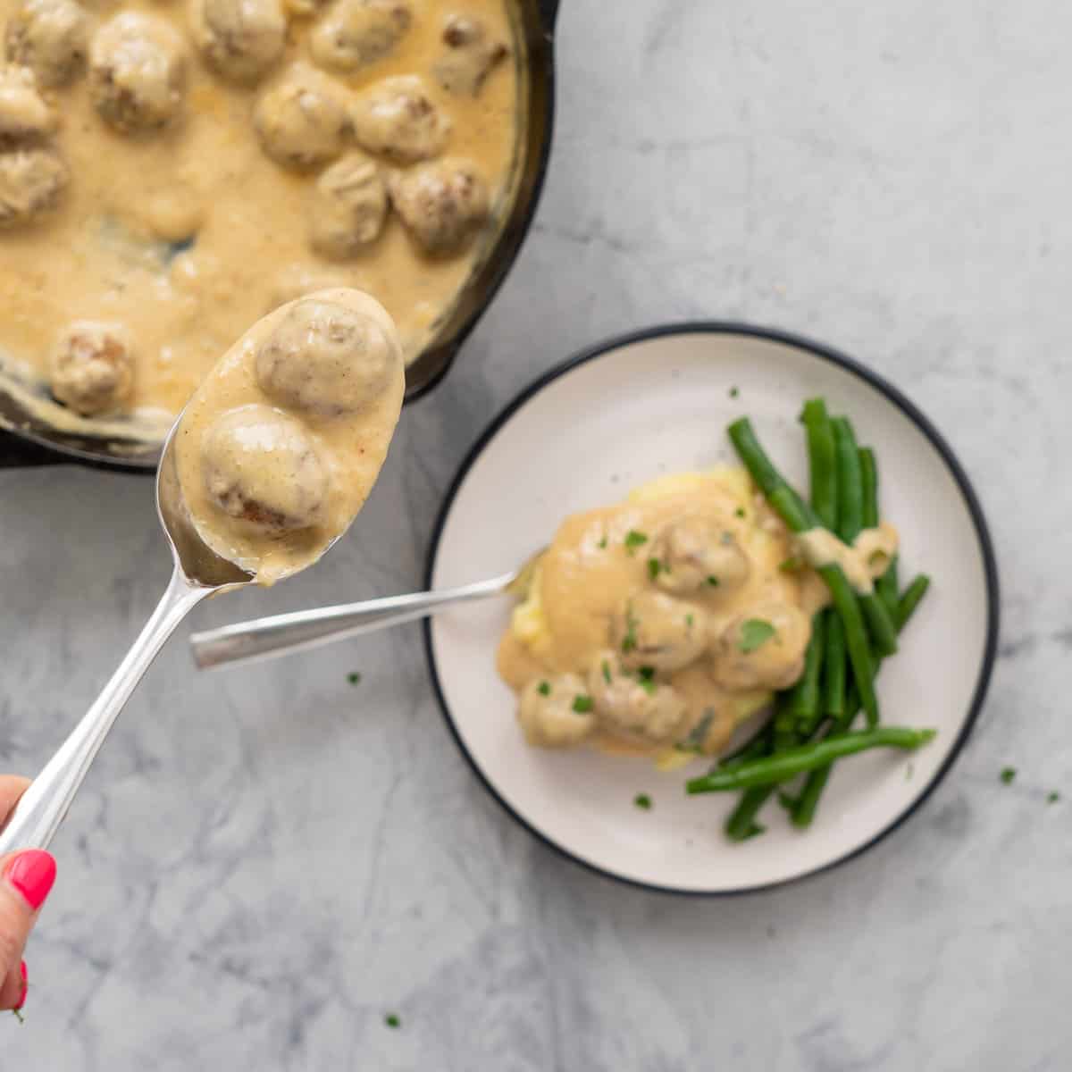 A spoonful of Sweedish meatballs raised above the rest of the batch below in a cast-iron pan next a plate with a serving over the top of mashed potatoes and side of blanched beans and a sprinkling of parsley.