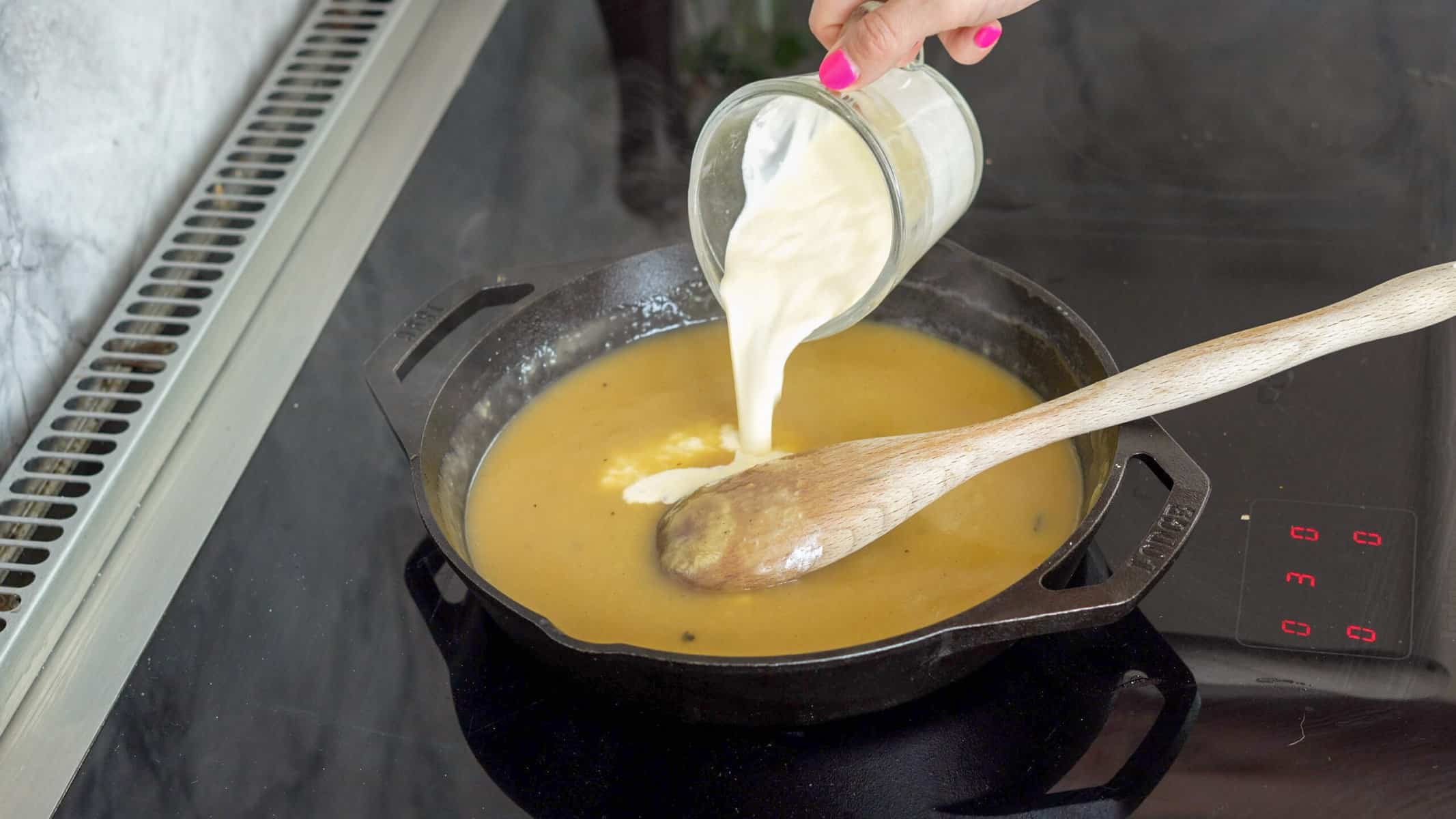 A small jug of cream being poured into the cast iron pan of melted butter by a wooden spoon.