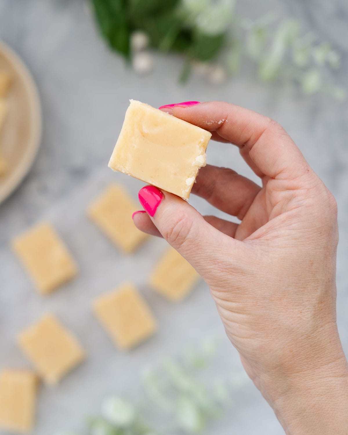 A hand with pink finger nails holding up a slice of vanilla fudge above the sliced batch below
