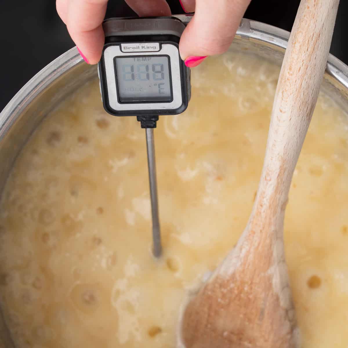 Fudge mixture brought to a bubbling in a saucepan on the bench element with a wooden spoon resting on the side and a thermometer taking the temperature
