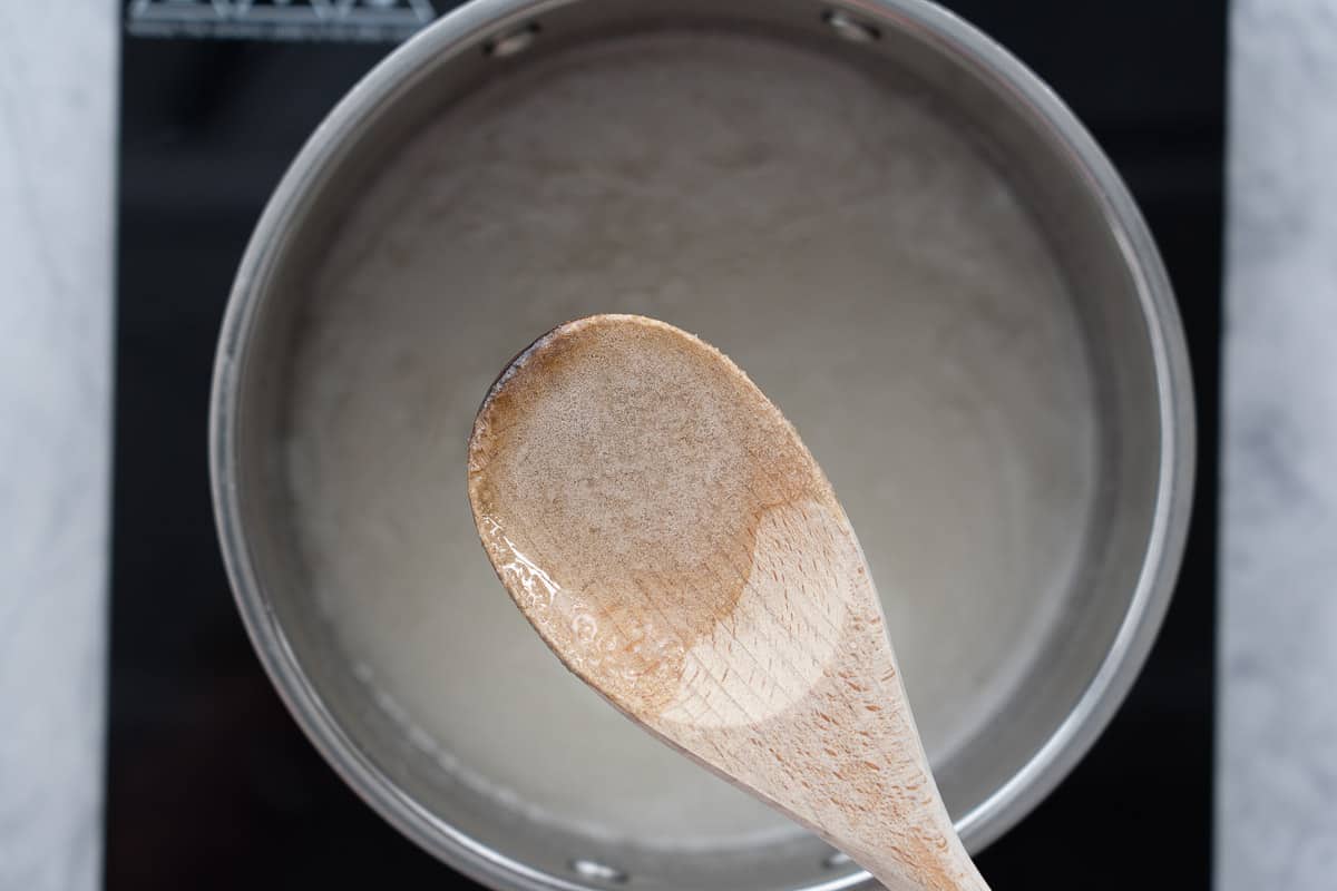A saucepan of dissolved sugar and water on sitting on a bench element with a wooden spoon raised above it showing the dissolved sugar crystals 