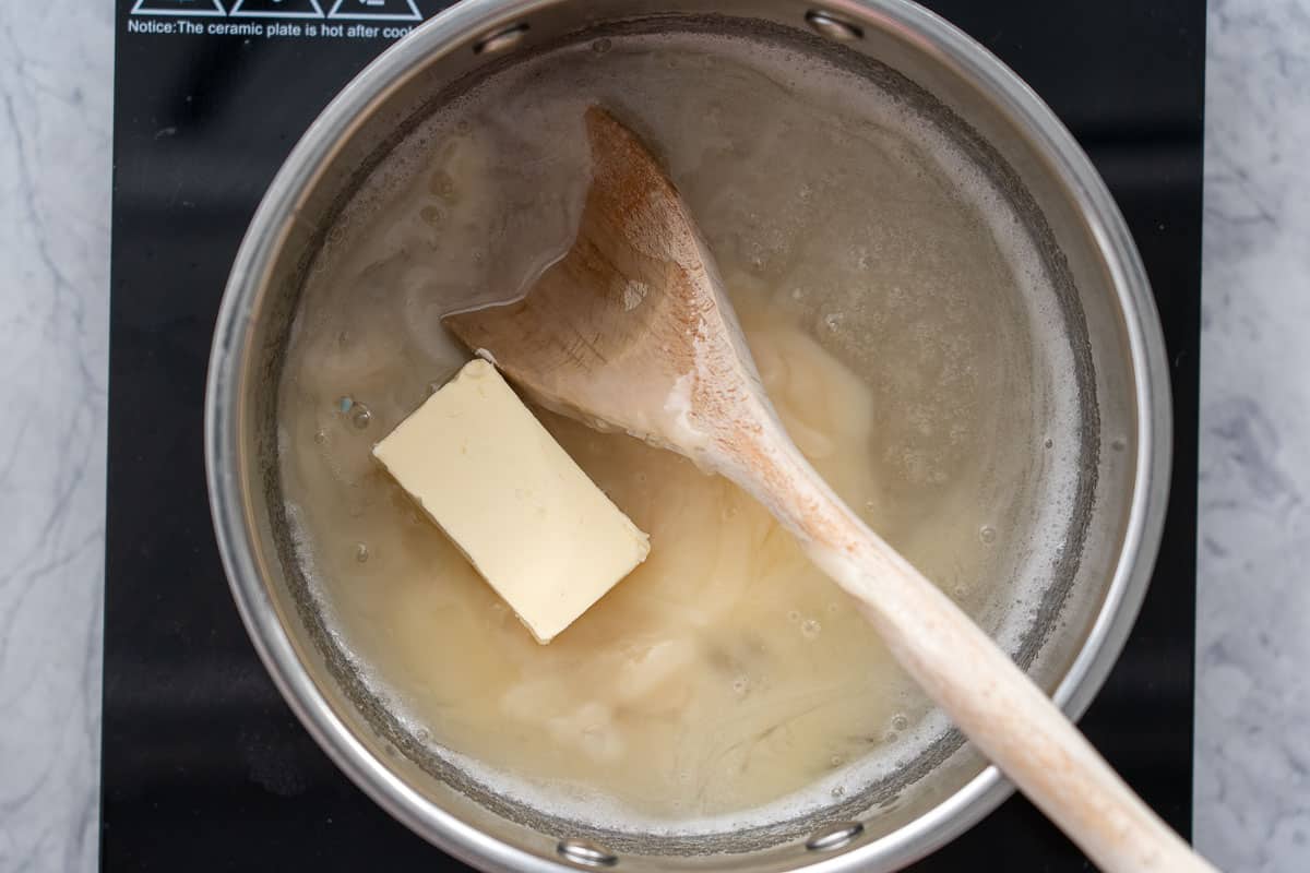 A saucepan of dissolved sugar and water with the butter added sitting on a bench element with a wooden spoon resting against the side. 