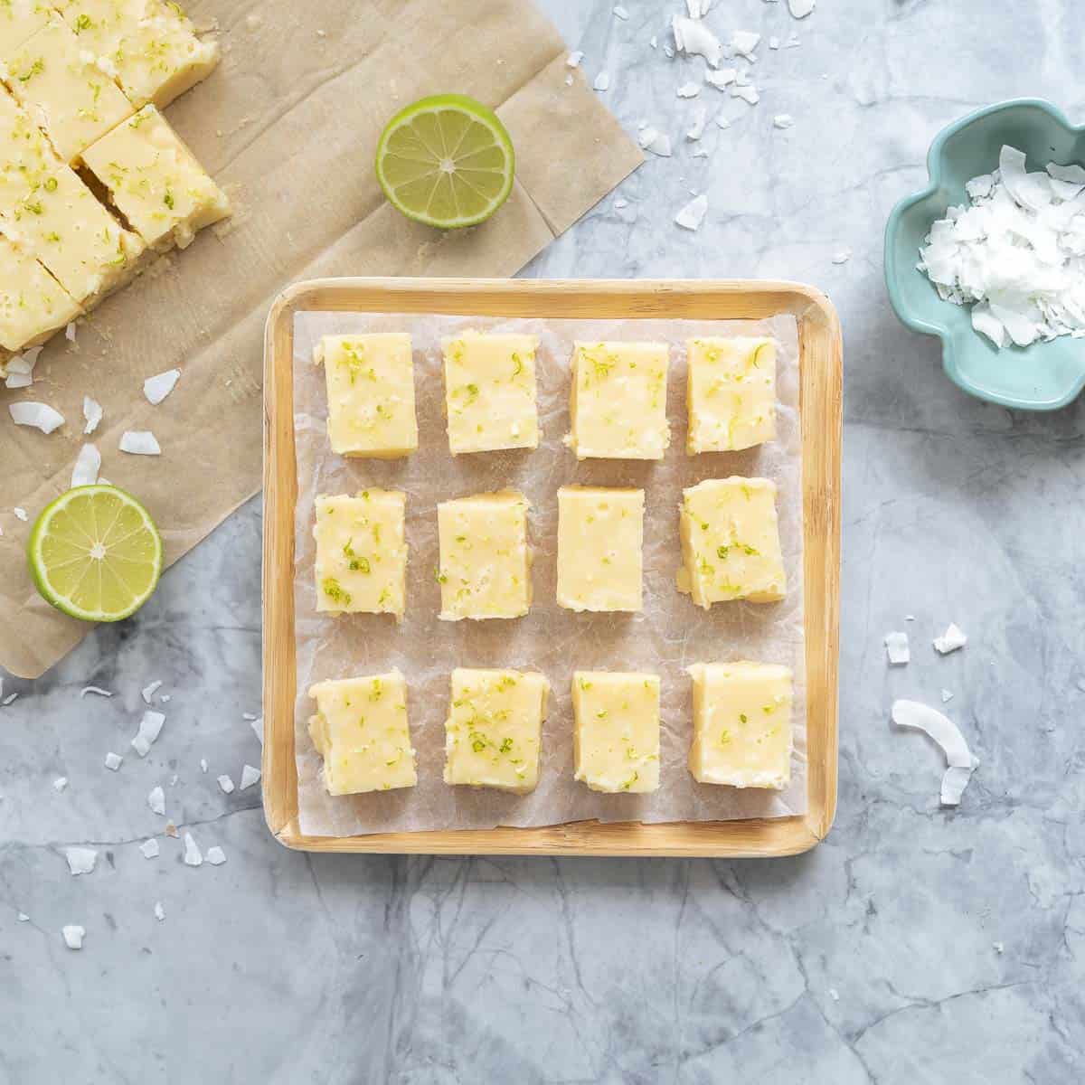 Slices of vegan fudge laid out on a piece of crinkled baking paper on a square wooden serving board next to a small serving dish of coconut flakes and halved limes and scattered coconut flakes on the bench