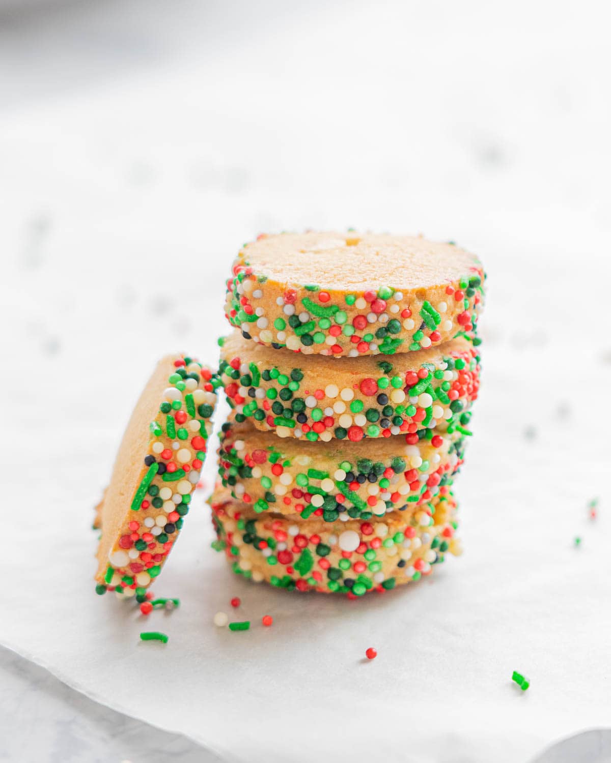 A stack of Christmas Sprinkle Cookies resting on the bench with one resting on its side against the stack. 