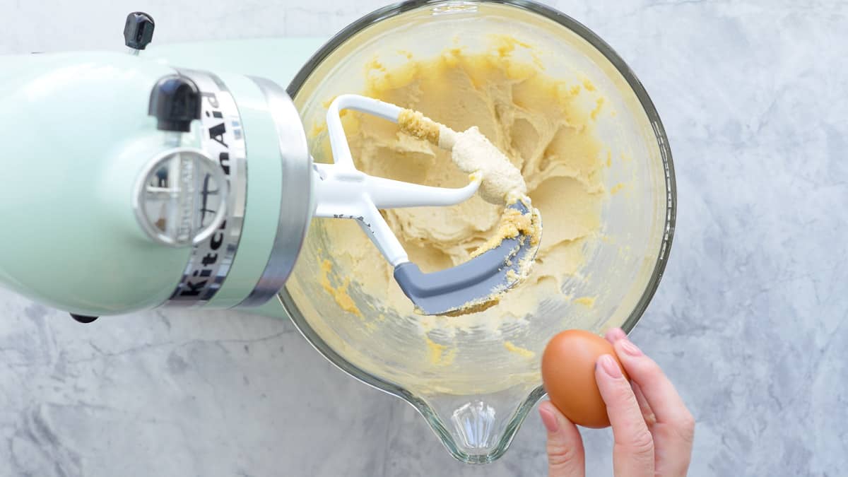 Creamed butter and brown sugar in a stand up mixer sitting on the bench with a hand holding an egg, about to crack it on the side of the glass bowl  