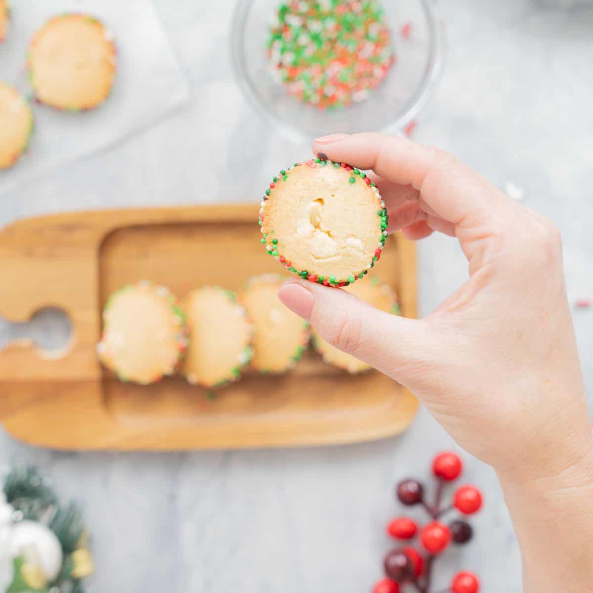A hand holding up a baked Christmas Sprinkle Cookie above a wooden board full of cookies with a couple of Christmas decorations scattered around it. 