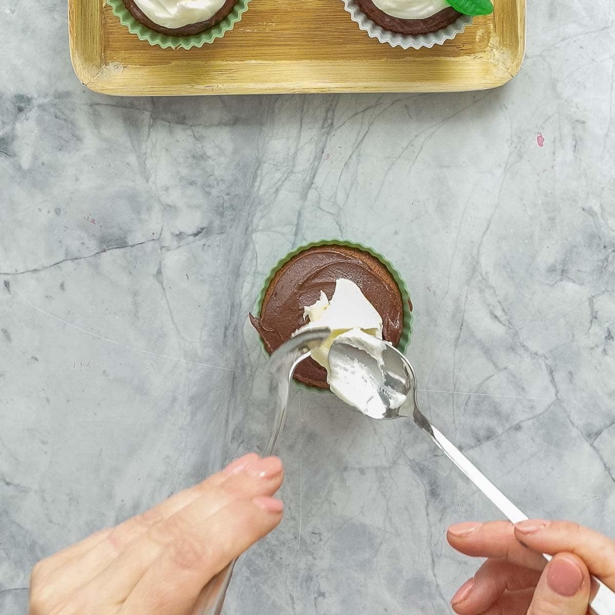 A spoonful of cream cheese frosting being placed on top of a chocolate frosted cupcake. 