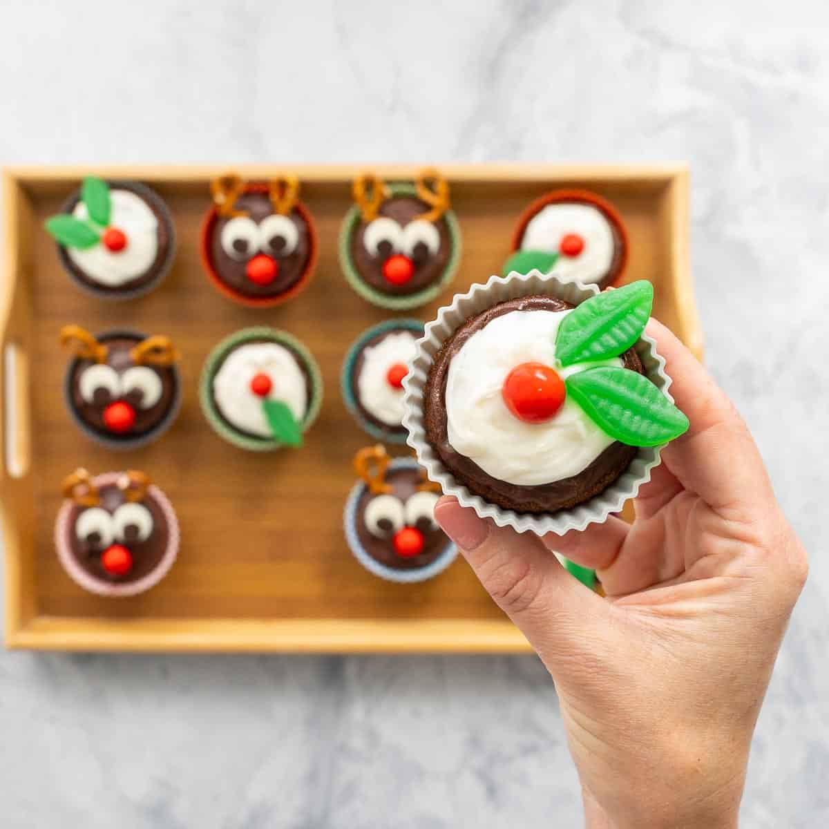 A cupcake decorated to look like a figgy pudding with frosting and candy being held up to the camera above a tray of cupcakes decorated to look like reindeer. 