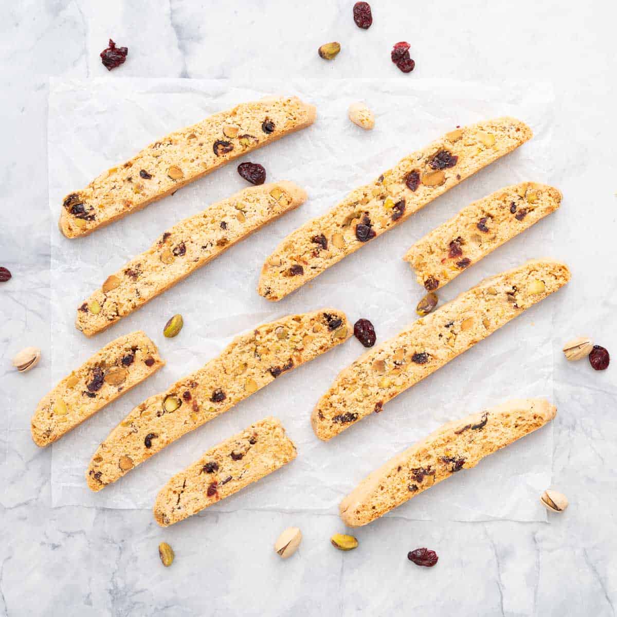 Pieces of biscotti studded with fruit and nuts on a crinkled piece of white parchment paper scattered with additional pistachios and cranberries
