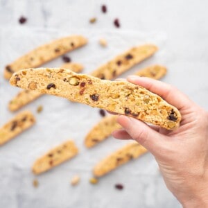 A piece of biscotti studded with pistachios and cranberries being held up to the camera.