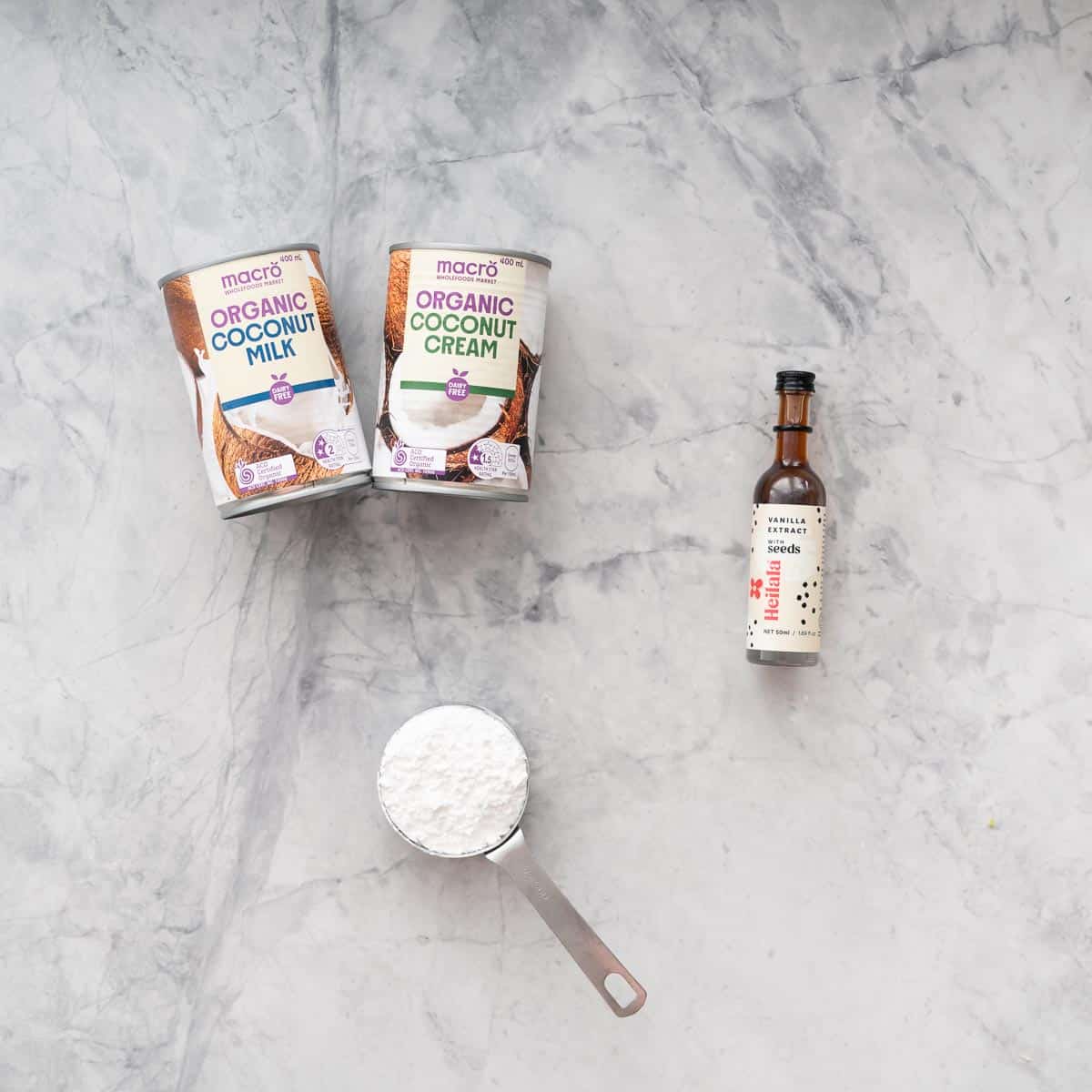 Canned coconut milk, a bottle of vanilla extract and icing sugar on a grey marble bench top.