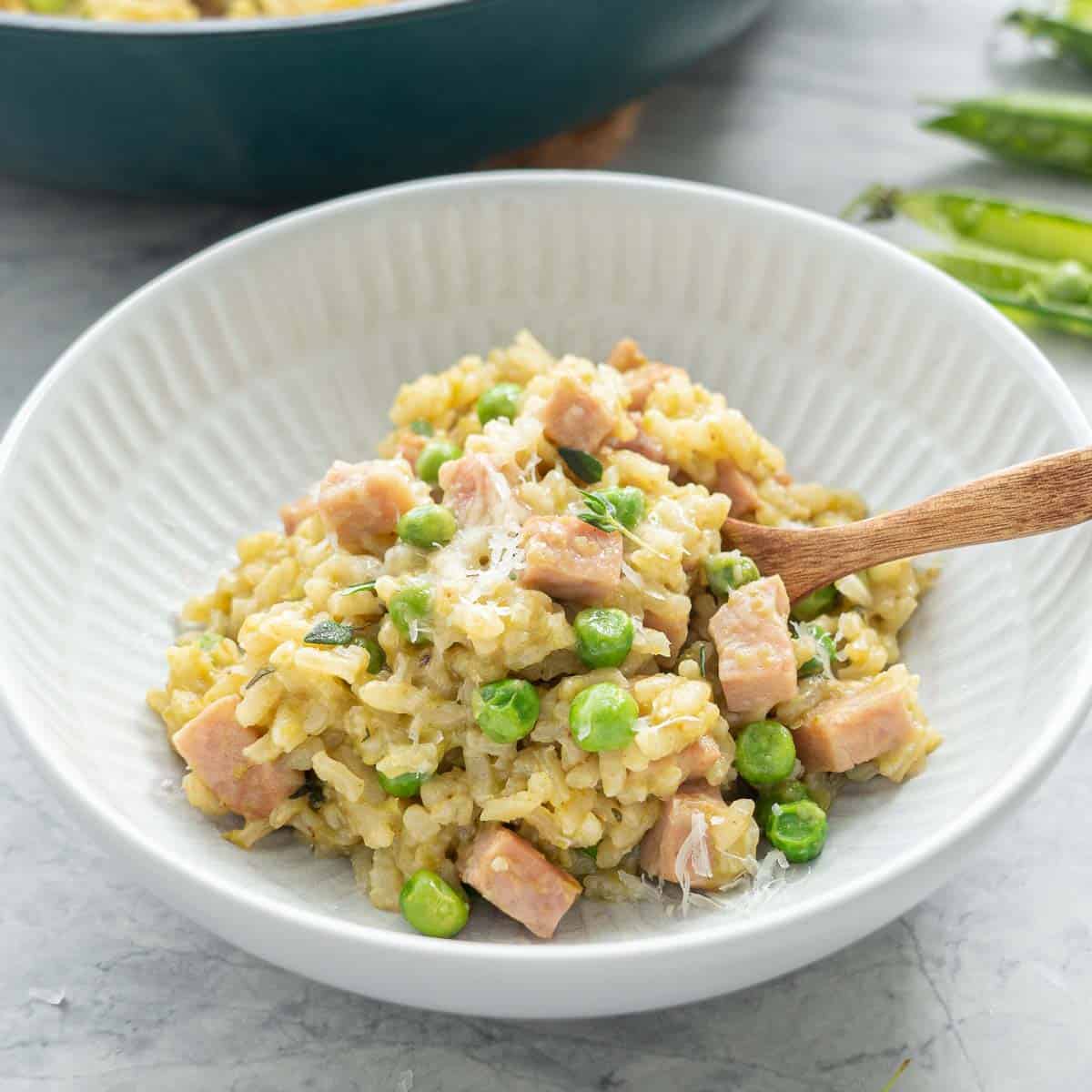 A bowl of kakhki green risotto studded with green peas and chunks of pink ham in a ceramic bowl with wooden spoon. 