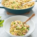 A bowl of kakhki green risotto studded with green peas and chunks of pink ham in a ceramic bowl with wooden spoon.