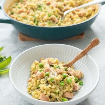 A bowl of kakhki green risotto studded with green peas and chunks of pink ham in a ceramic bowl with wooden spoon.