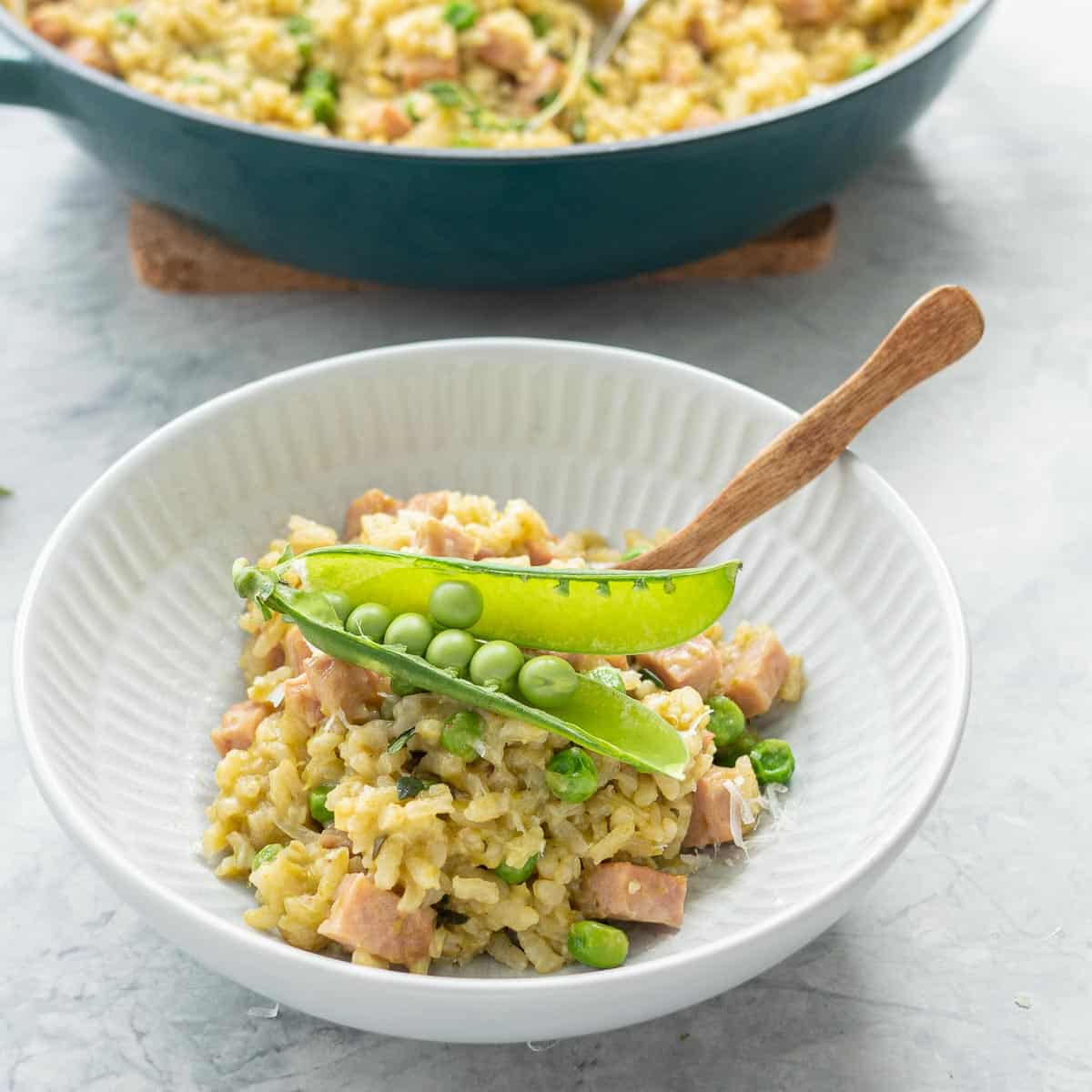 A bowl of kakhki green risotto studded with green peas and chunks of pink ham in a ceramic bowl with wooden spoon topped with a pea pod