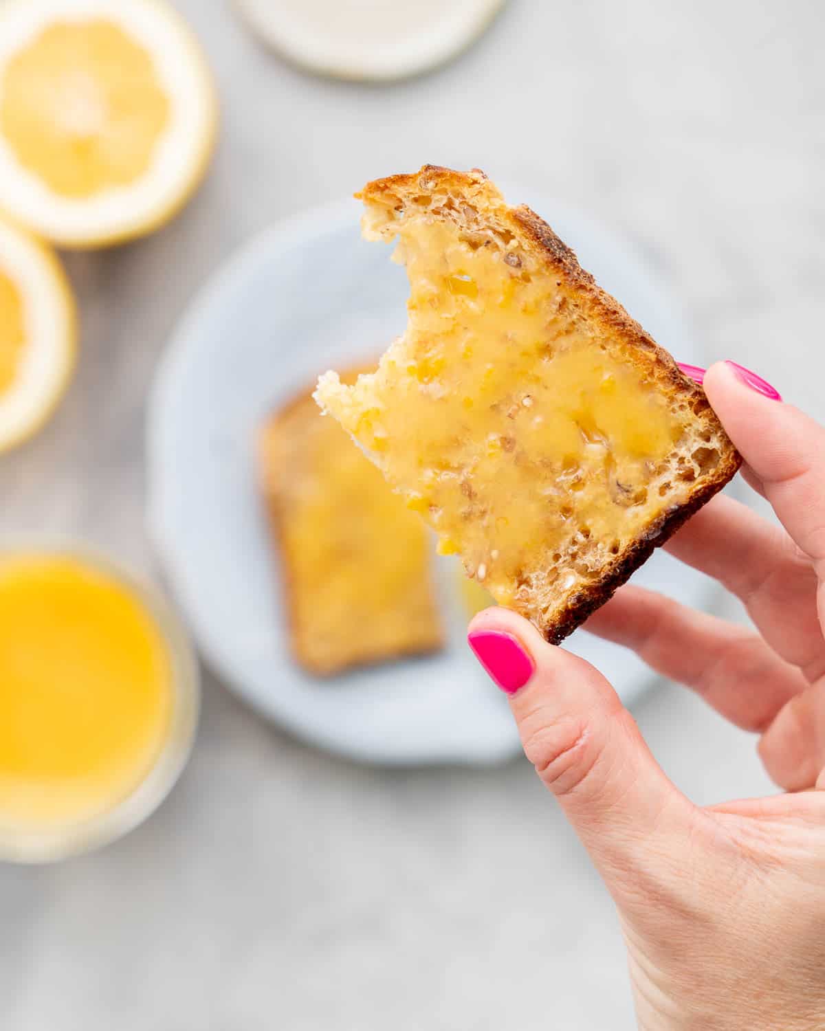 A hand holding up a slice of toast with Lemon Butter spread on it which is held above the other half of toast on a plate below