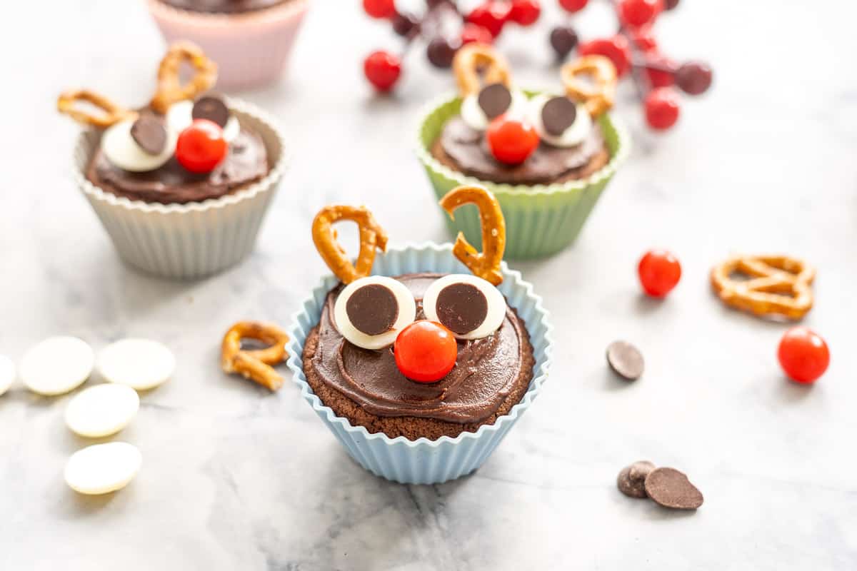 A cupcake decorated like a reindeer in a blue cupcake liner on a bench top scattered with candy. pretzels and chocolate drops. 