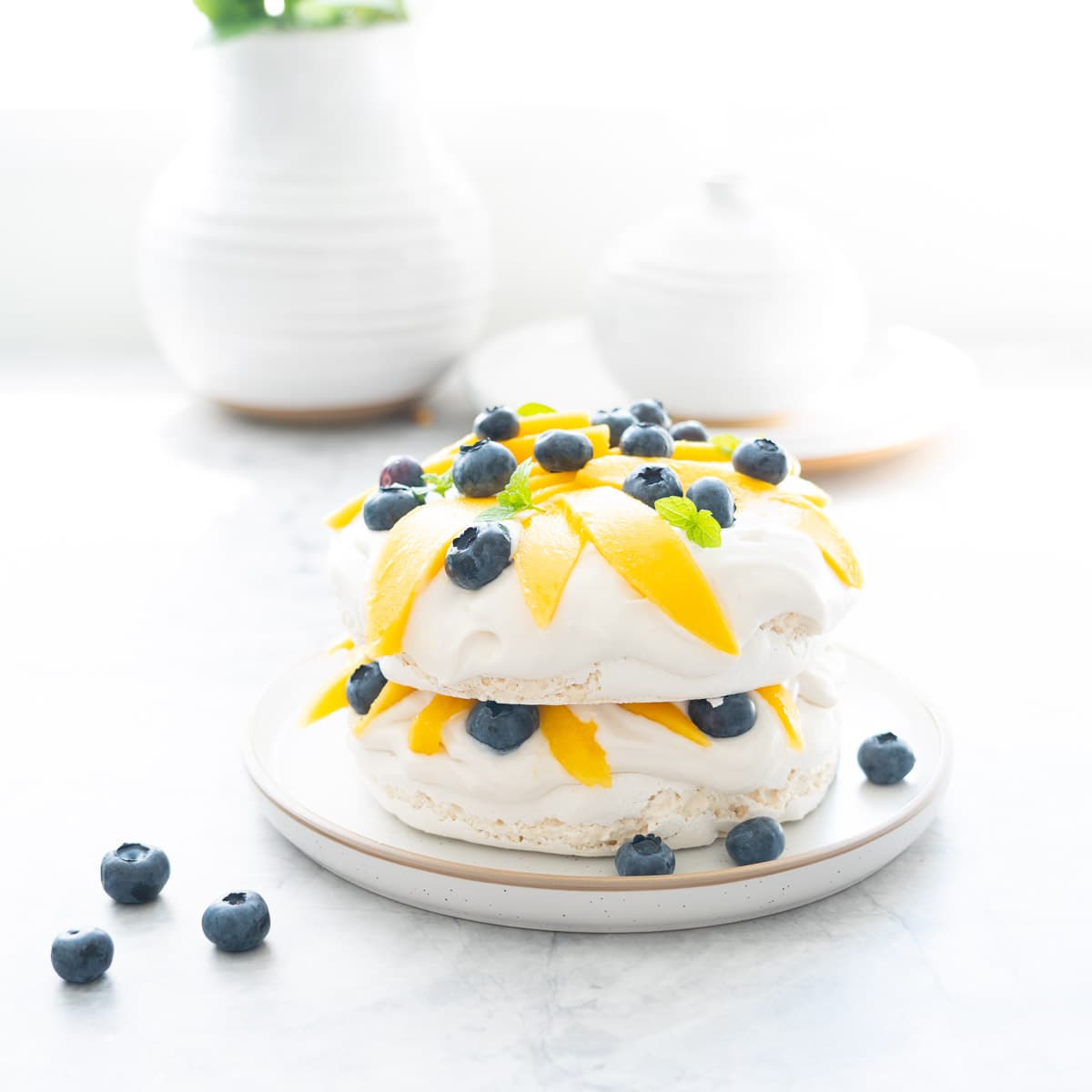 A two layer pavlova topped and filled with whipped cream, sliced mango, blueberries and mint leaves