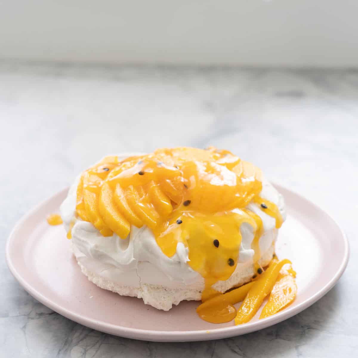A pavlova on a pink plate topped with whipped cream, sliced mango and passionfruit pulp.  