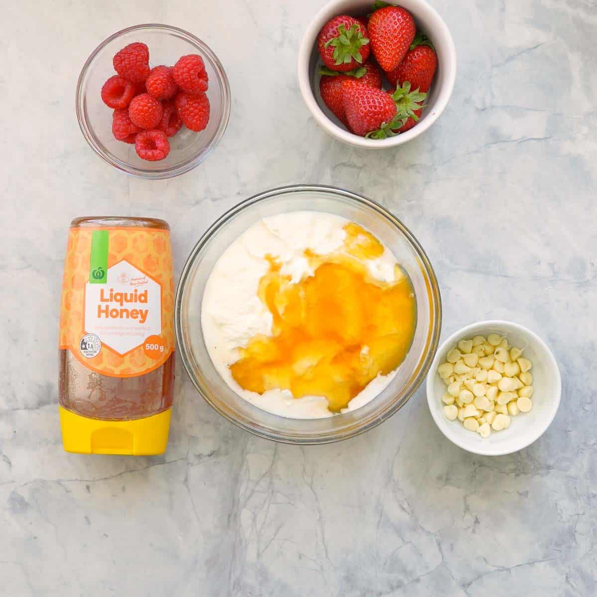 A small glass mixing bowl with yogurt, vanilla extract and honey sitting next to a bottle of liquid honey and  ramekins of raspberries, strawberries and white chocolate drops on the bench