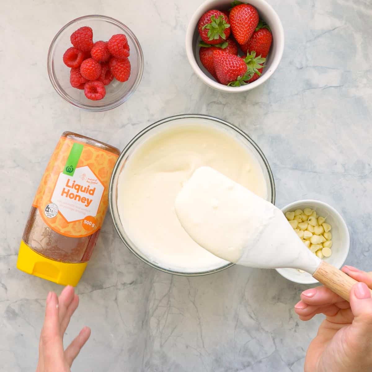 A small glass mixing bowl with yogurt, vanilla extract and honey stirred together with a spatula which issitting next to a bottle of liquid honey and  ramekins of raspberries, strawberries and white chocolate drops on the bench