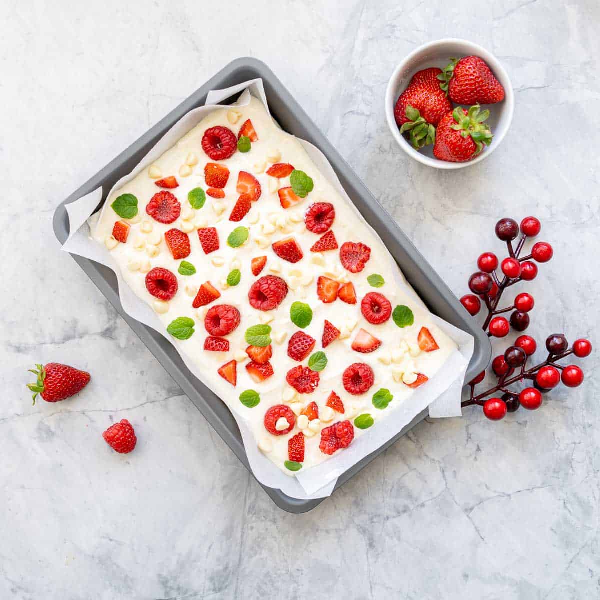 A lined tray of yoghurt bark which is scattered with sliced strawberries, raspberries and baby mint leaves scattered over the top which is sitting next to a small ramekin of strawberries and a Christmas holly decoration