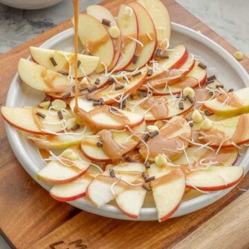 Caramel sauce being poured over a platter of apple slices, chocolate chips and shredded coconut.