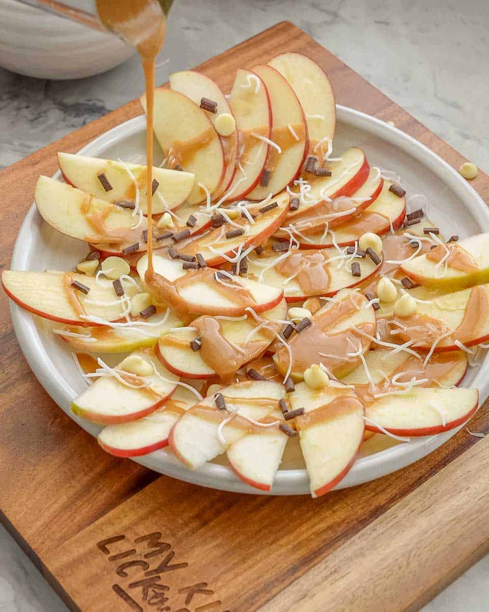 Caramel sauce being poured over a platter of apple slices, chocolate chips and shredded coconut.