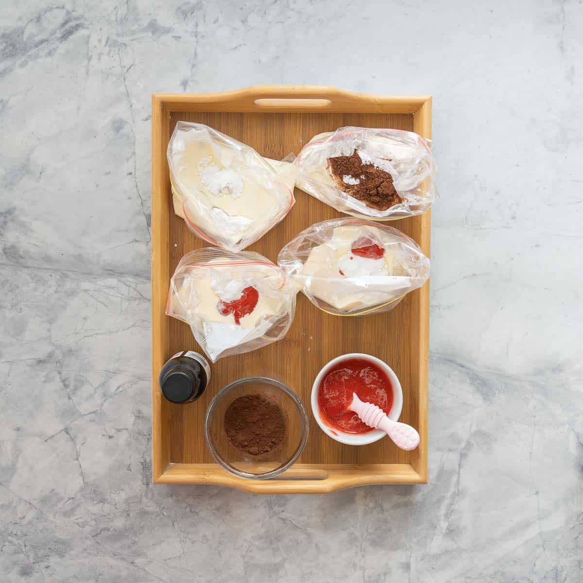 Three zip lock bags of cream on a wooden tray with small bowls of strawberry puree, cocoa and vanilla extract.