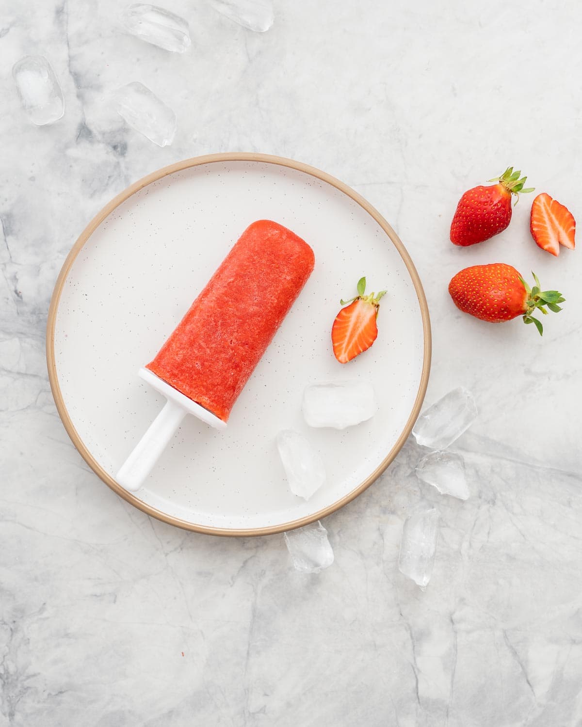 A strawberry popsicle resting on a white plate scattered with ice cubes and pieces of strawberries.
