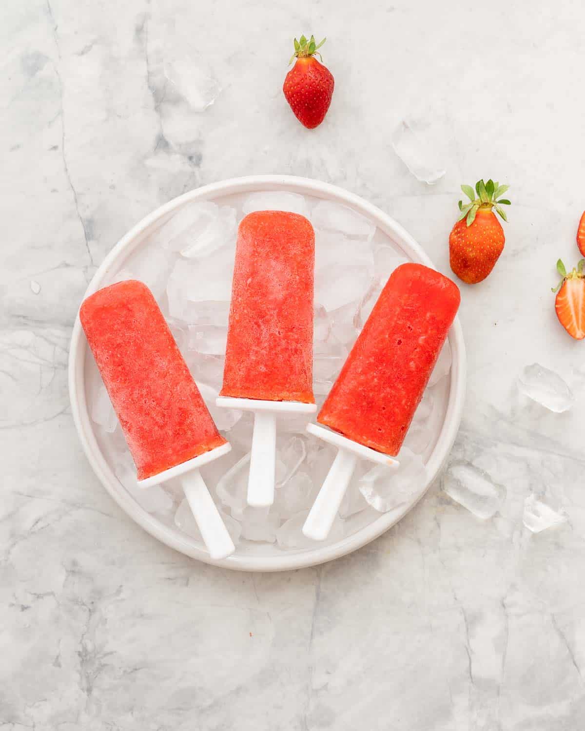 Three red fruit popsicles resting on a bowl filled with ice cubes. 