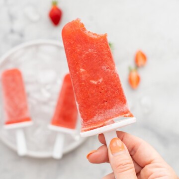 A strawberry popsicle with one bite taken out of it being held up to the camera.