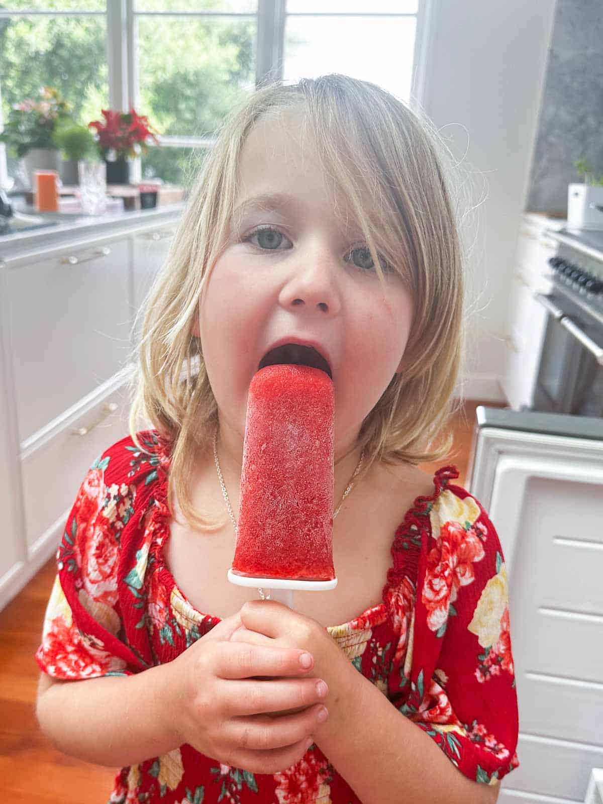 A young girl in a red floral dress liking a strawberry popsicle. 