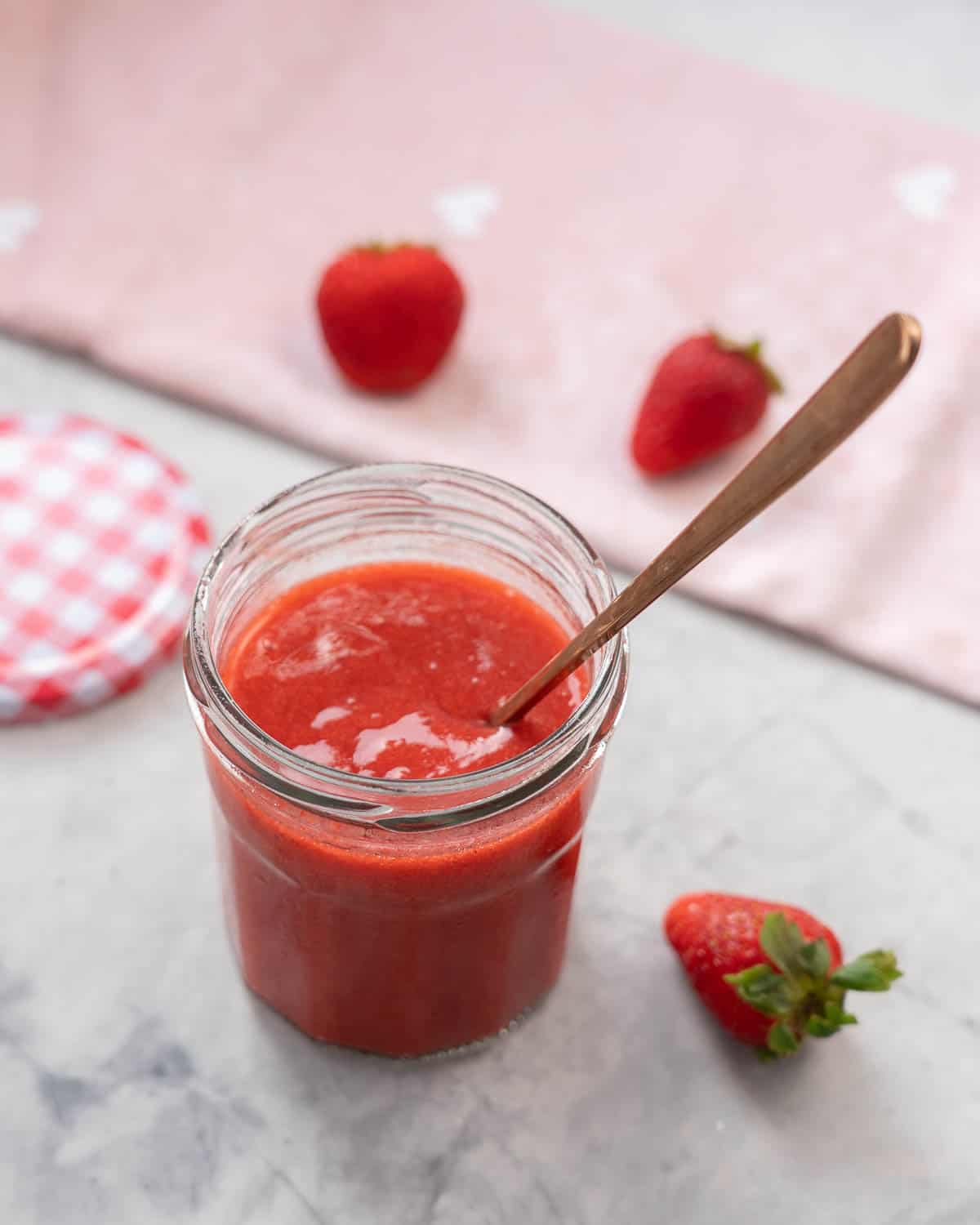 A glass jar of red fruit puree with a golden spoon resting in it, strawberries scattered on the bench around the jar. 