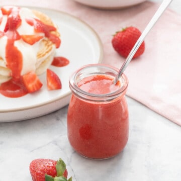 A small glass jar of strawberry puree in front of a plate of pancakes drizzled with the syrup.