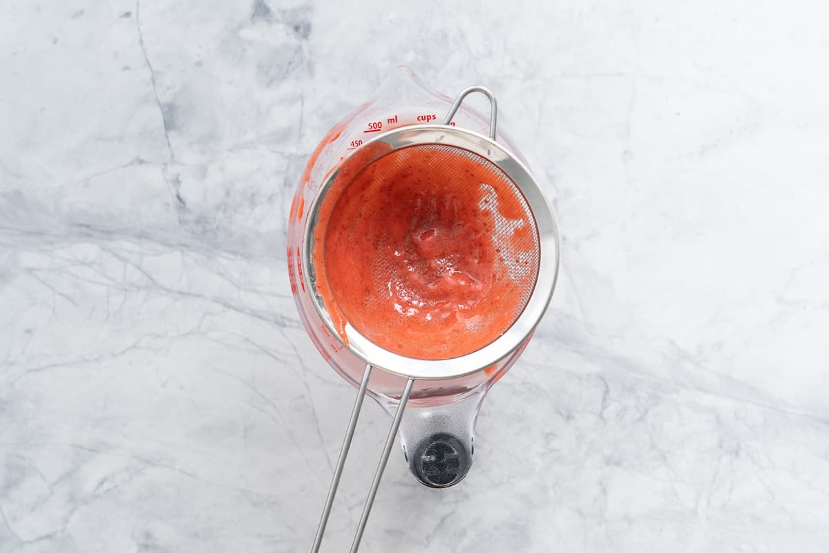 Strawberry puree being strained through a sieve into a glass measuring jug. 
