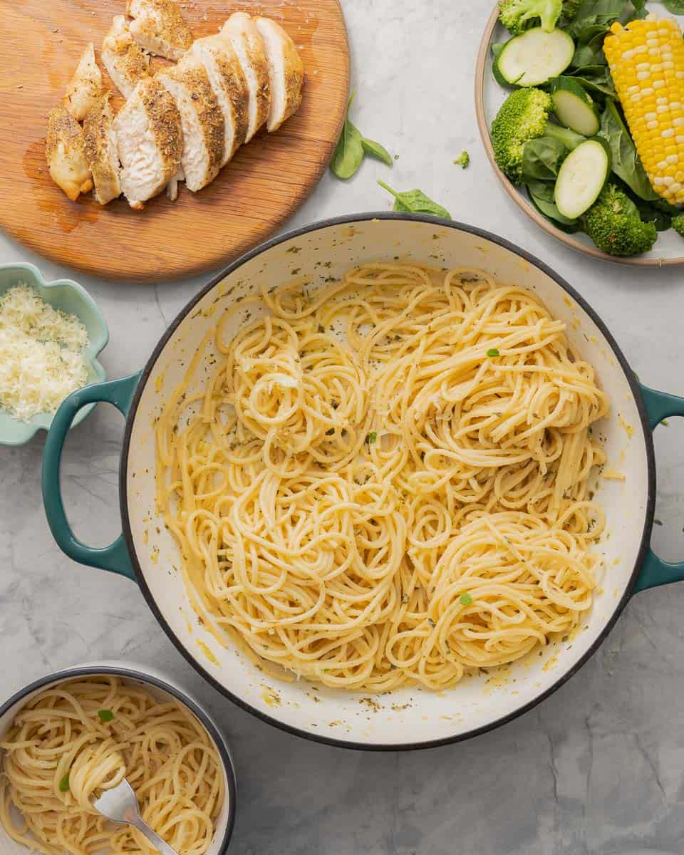 A large pan full of buttered noodles resting on the bench next to a cooked and sliced chicken breast and a plate of vegetables