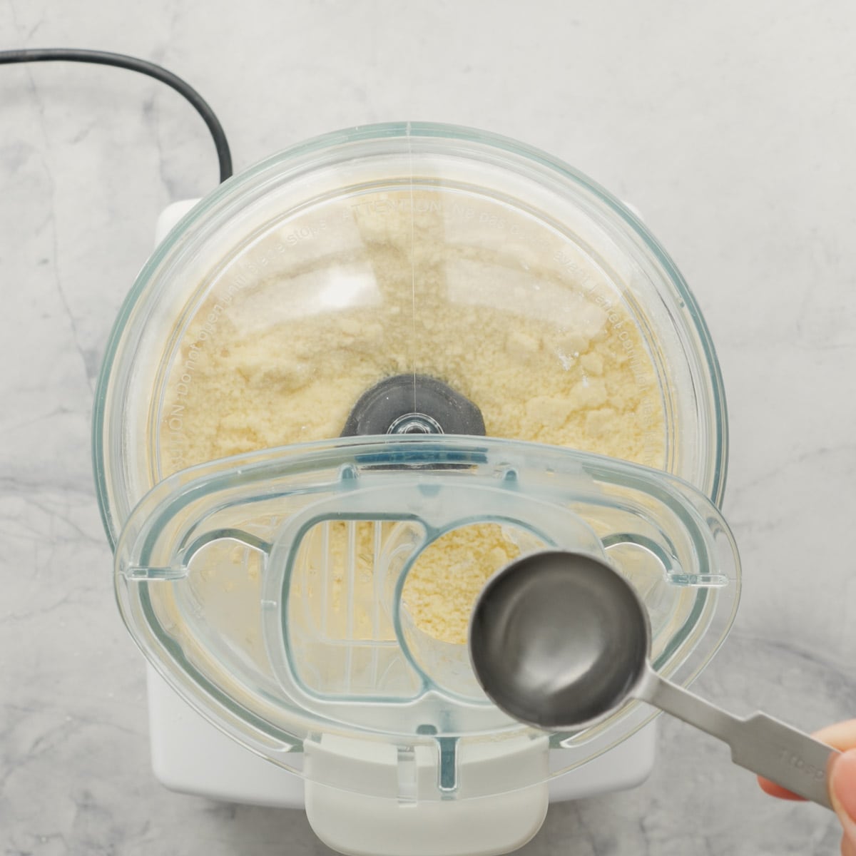 A food processor sitting on the bench with the lid on and a table spoon of water above it, held by a hand.