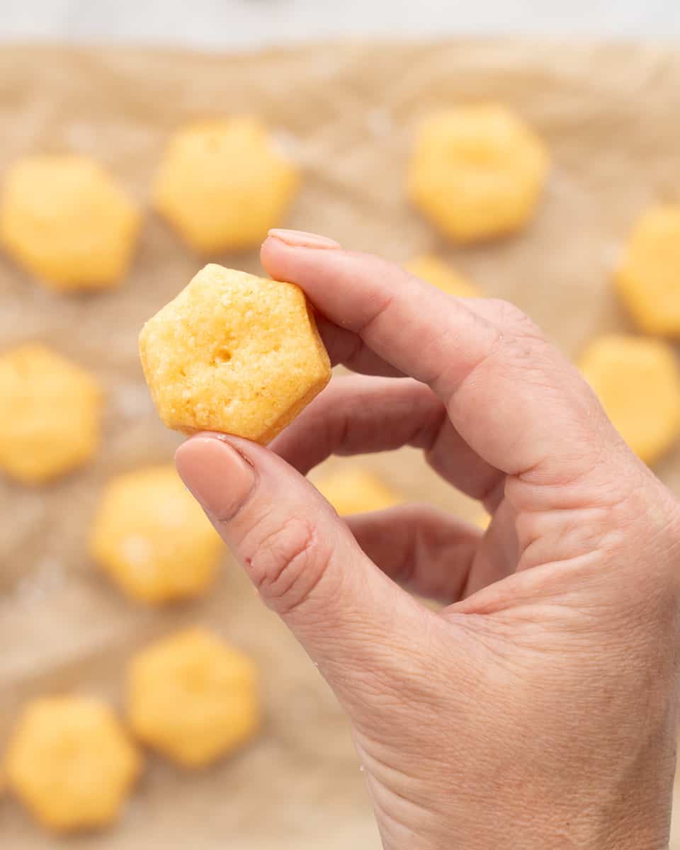 A hand holding one cheese cracker above the batch of golden brown baked cheese crackers which are resting on lined baking tray. 