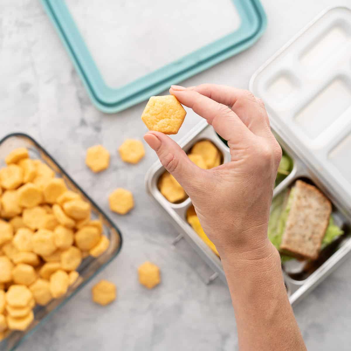 A hand holding one cheese cracker above the batch of golden brown baked cheese crackers in a airtight container and a tin lunch box 