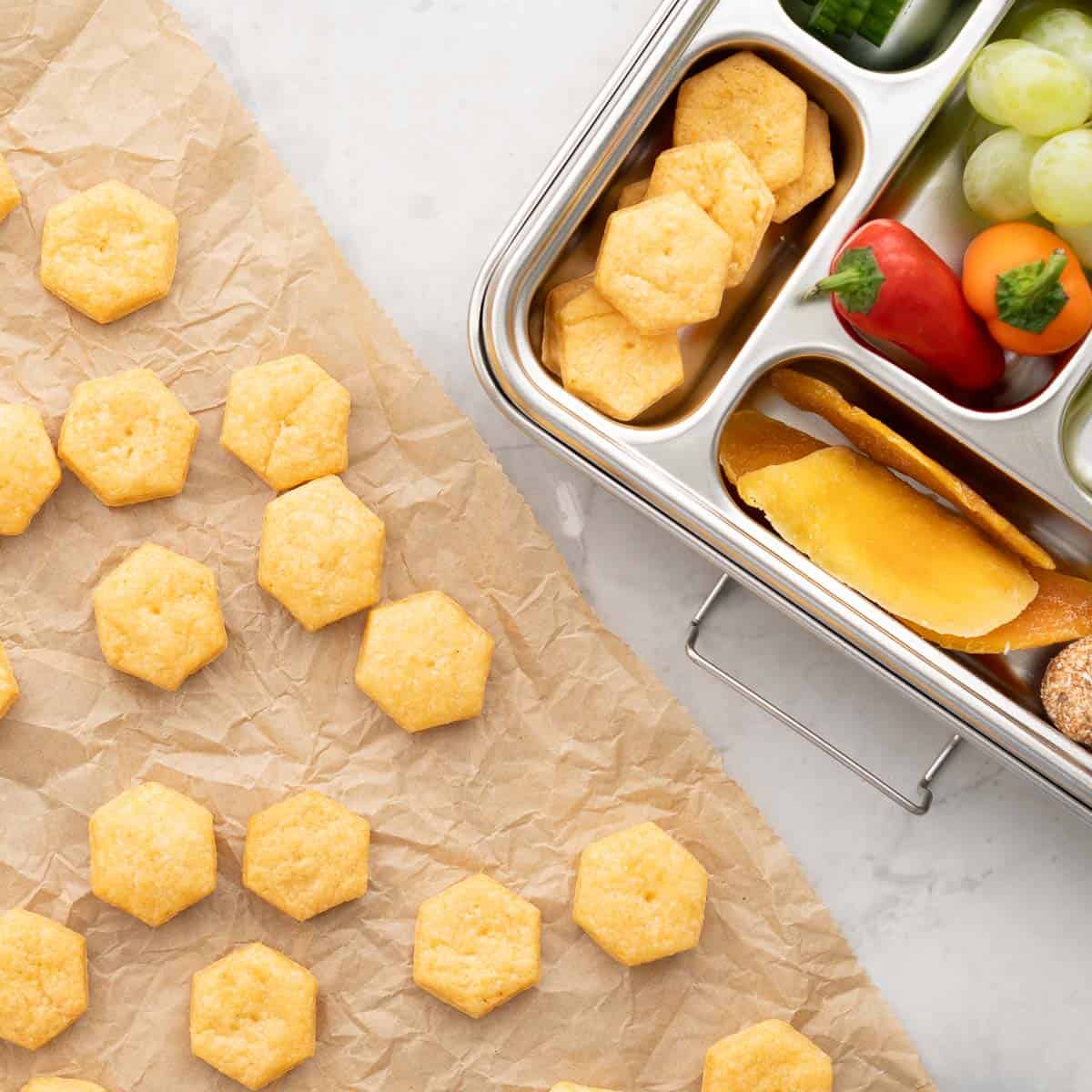Cheese crackers resting on brown baking paper on the bench next to a tin lunch box with individual compartments filled with dried mango, capsicums grapes and more of the cheese crackers 