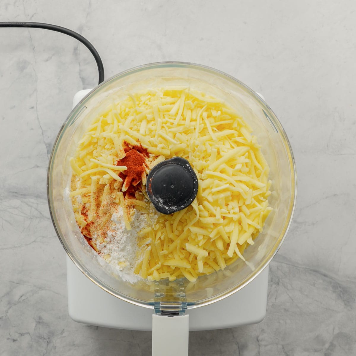 A food processor sitting on the bench with cheese, gluten free flour and seasoning inside it.