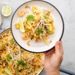 A white plate with black rim holding pad this, with bean sprouts, lime wedges and prawns visible above a large fry pan of pad thai.