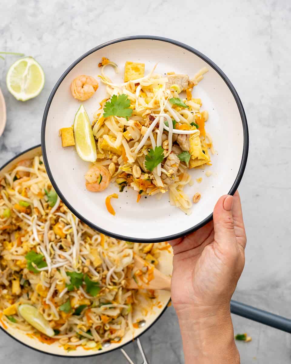 A white plate with black rim holding pad this, with bean sprouts, lime wedges and prawns visible above a large fry pan of pad thai.