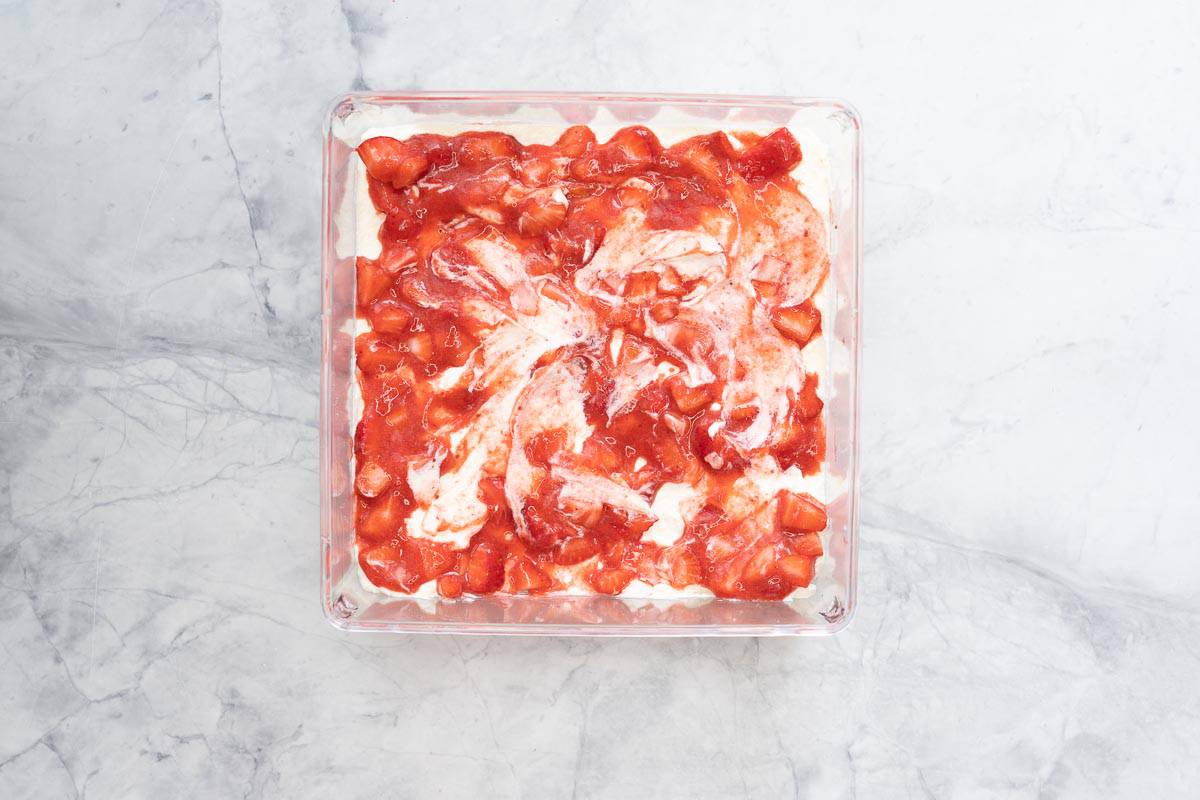 A square glass container of strawberries, strawberry puree and whipped cream. 